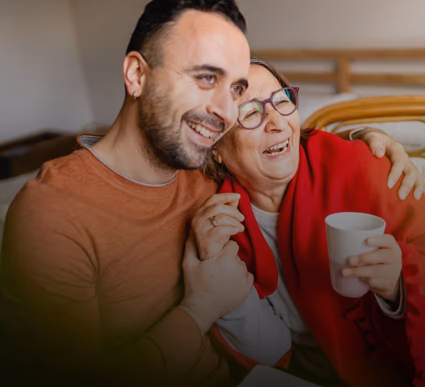 Smiling man hugging an elderly woman who is wearing glasses and a red shawl, holding a white mug while they sit close together indoors.