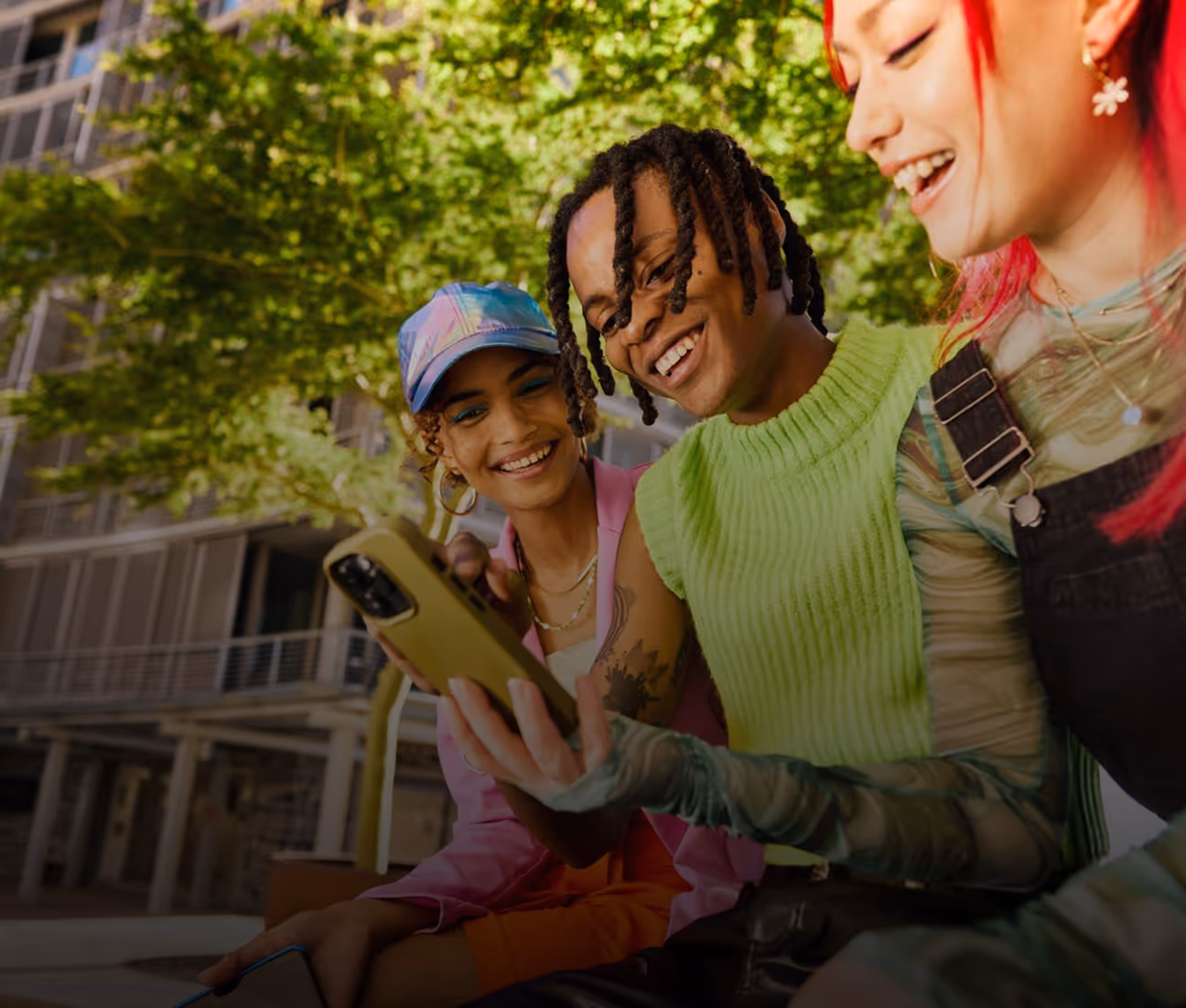Three young adults sitting outdoors on a bench, smiling and looking at a smartphone held by the person in the middle, with green trees and urban buildings in the background.