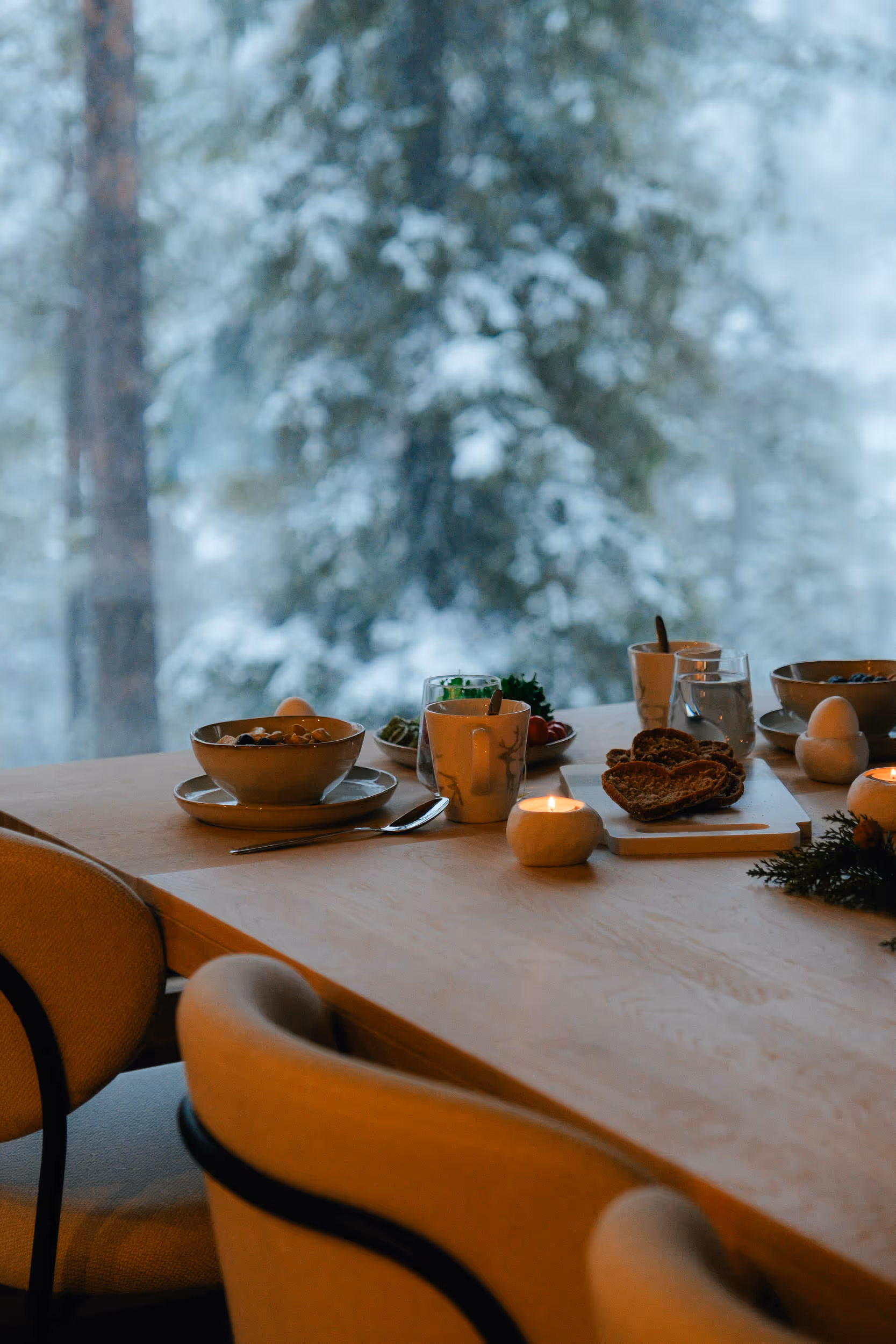 Cozy wooden table set for breakfast with bowls, mugs, candles, and bread, overlooking a snowy forest through a window.