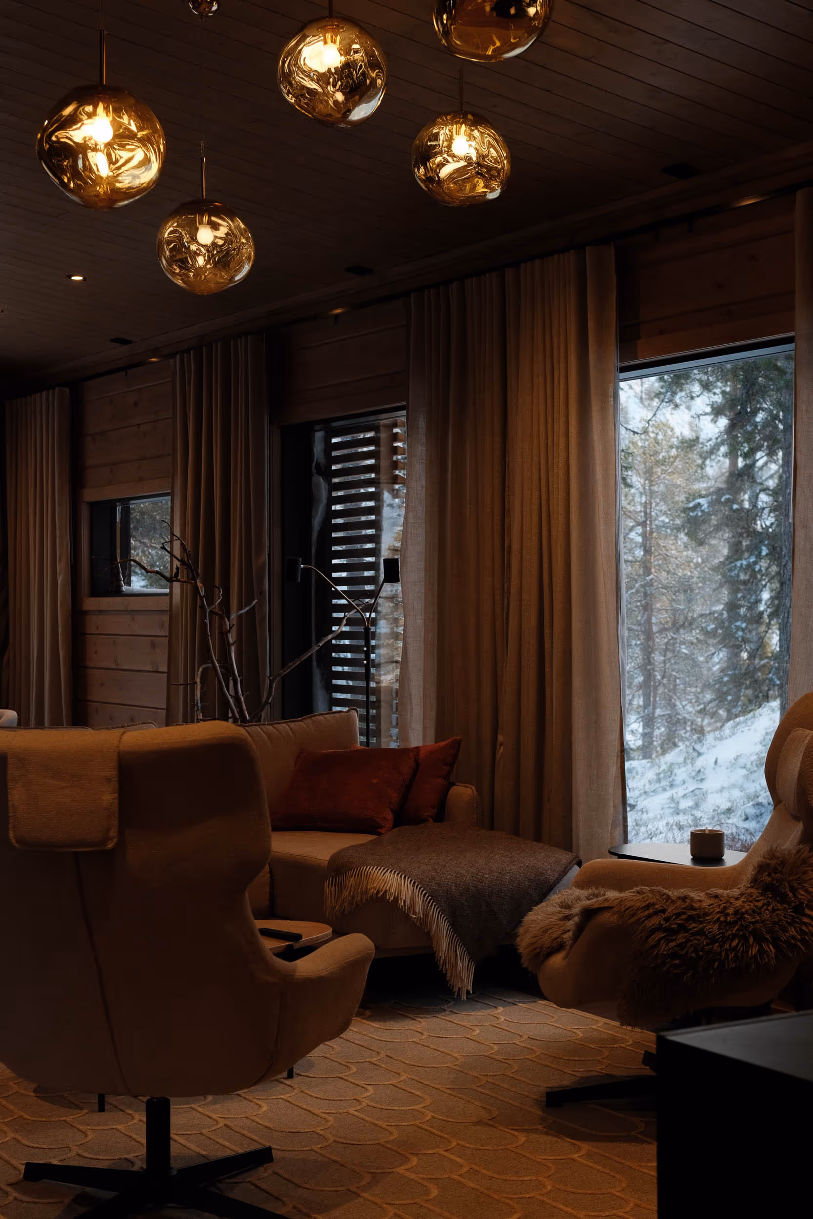 Cozy living room with beige armchairs, sofa with red pillows, a brown blanket, warm pendant lights, and snow-covered trees visible through large windows.