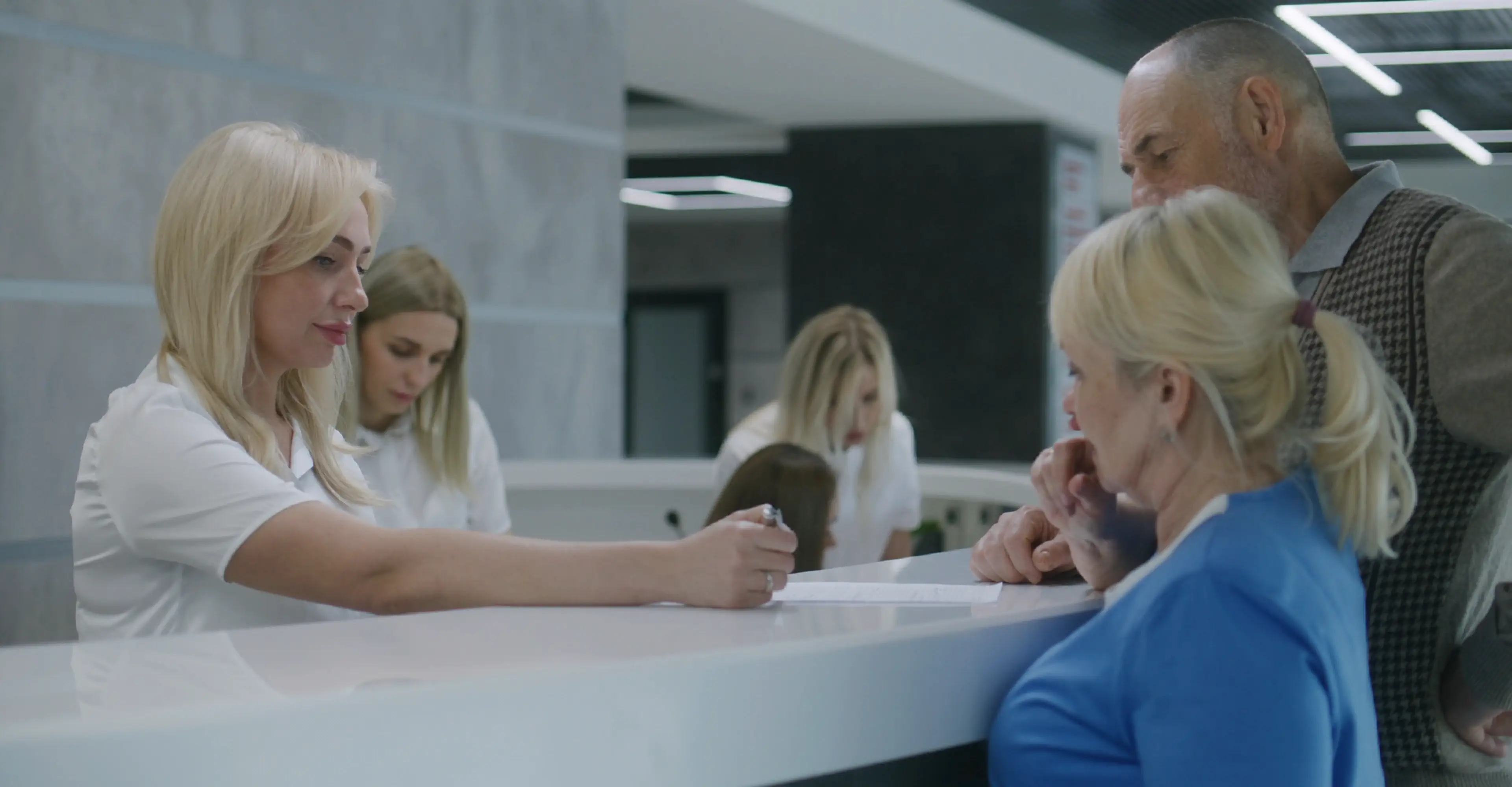 Blonde receptionist assisting elderly couple at a modern white counter in a brightly lit office or clinic.