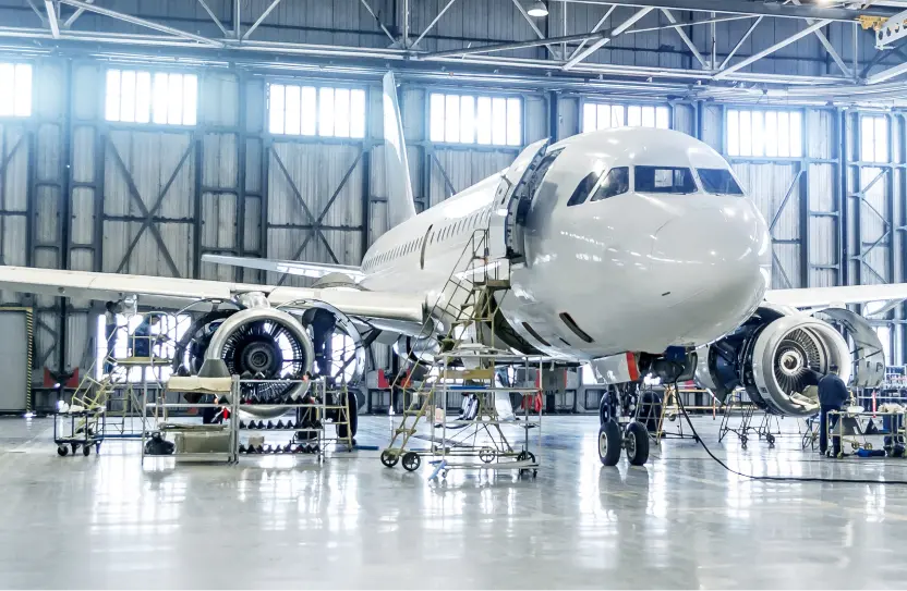 White commercial airplane inside a large hangar undergoing maintenance with several portable work platforms and technicians working on its engines and fuselage.