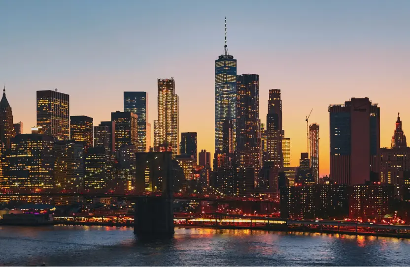 Evening view of a city skyline featuring illuminated skyscrapers and buildings reflecting on a river, with a bridge in the foreground under a clear sky.