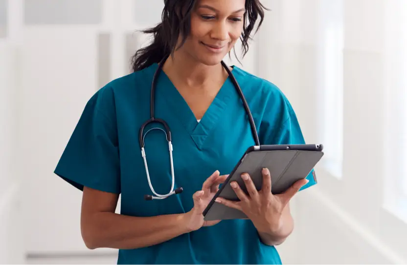 Female healthcare professional wearing teal scrubs and a stethoscope around her neck while using a tablet in a bright clinical setting.