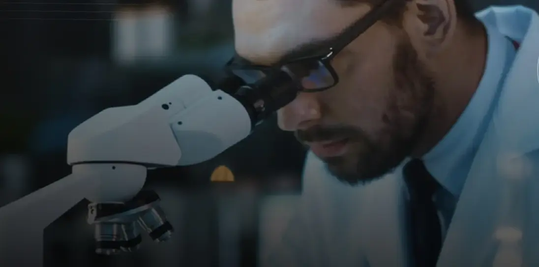 Man with glasses closely examining something through the eyepiece of a white microscope in a dimly lit laboratory or research setting.