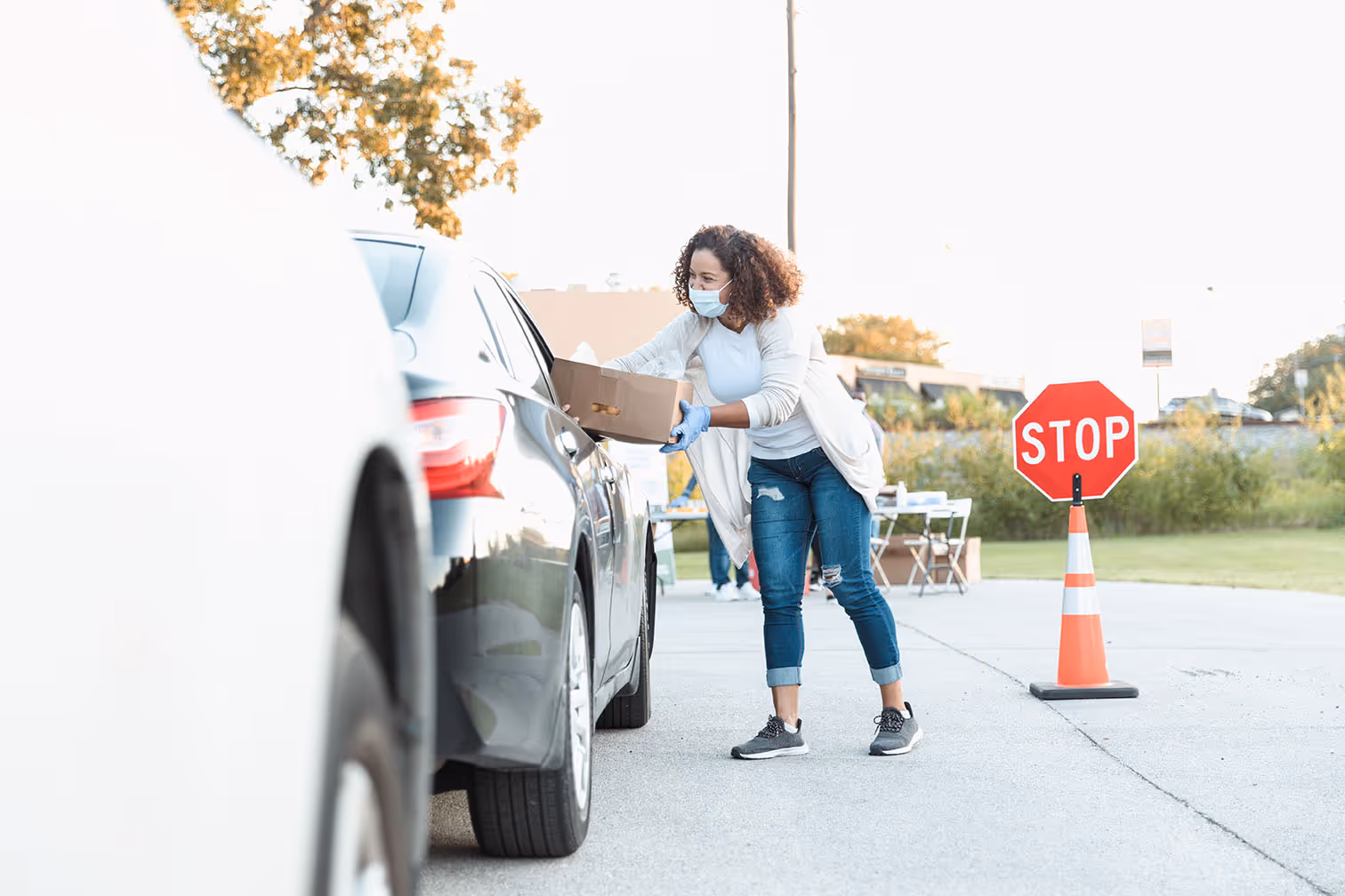 Woman wearing mask and gloves handing a cardboard box through the window of a black car at a drive-through distribution.