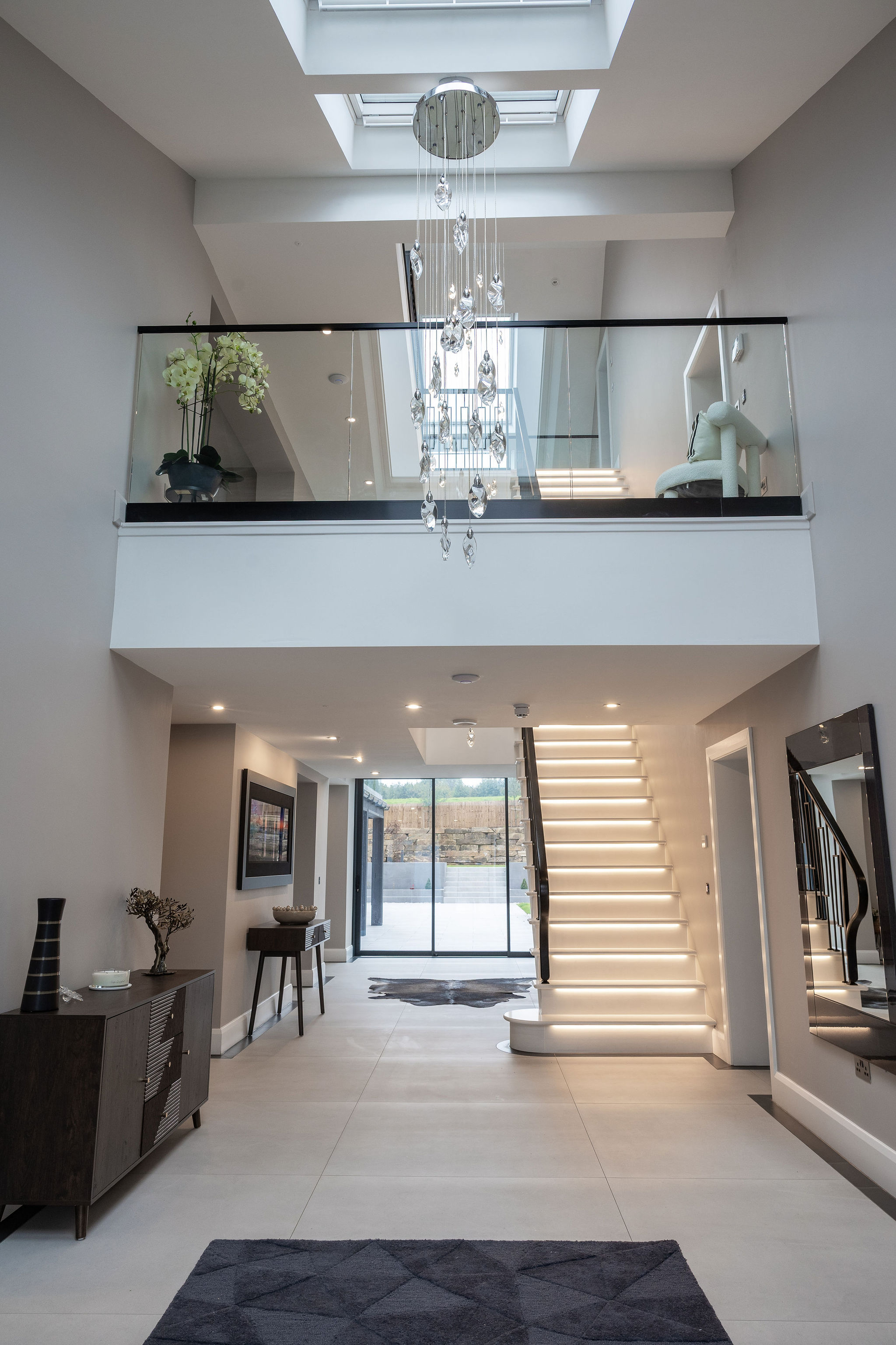 Modern, spacious hallway with illuminated staircase, glass balcony, hanging crystal light fixture, and sliding glass doors at the far end.