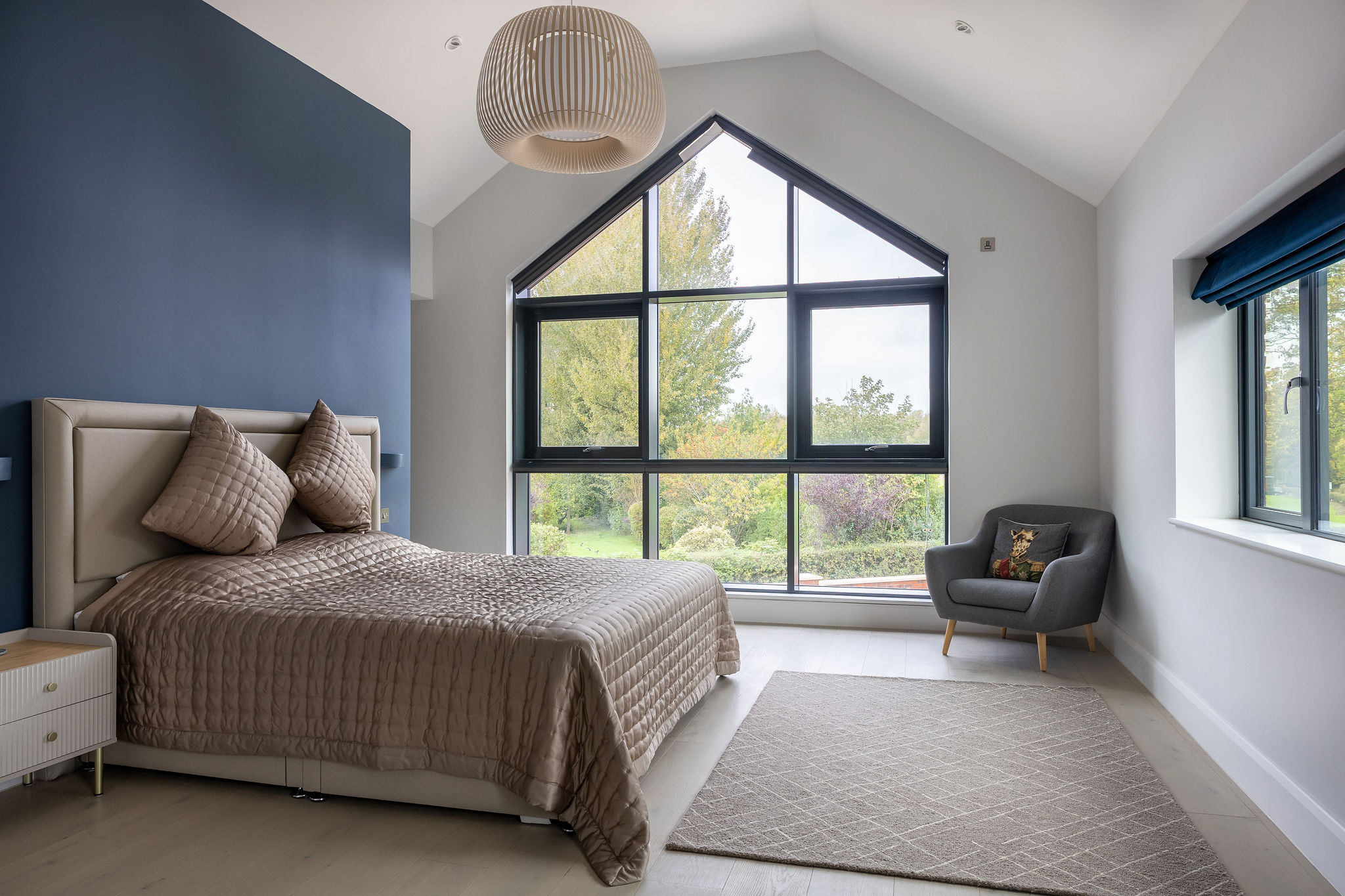Modern bedroom with large triangular window overlooking greenery, bed with beige quilted bedding, and gray armchair with decorative pillow.
