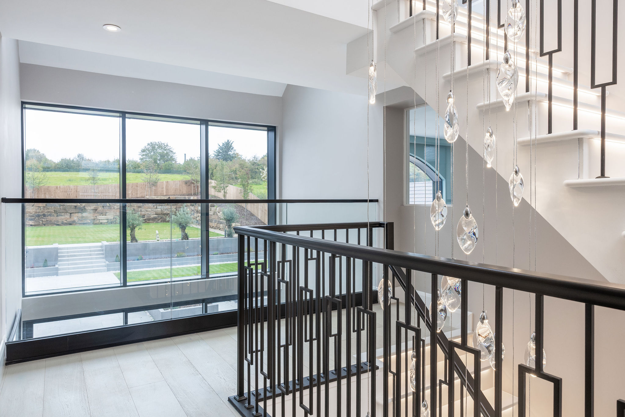 Modern staircase with black metal railings, hanging glass pendant lights, and large windows overlooking a green garden.