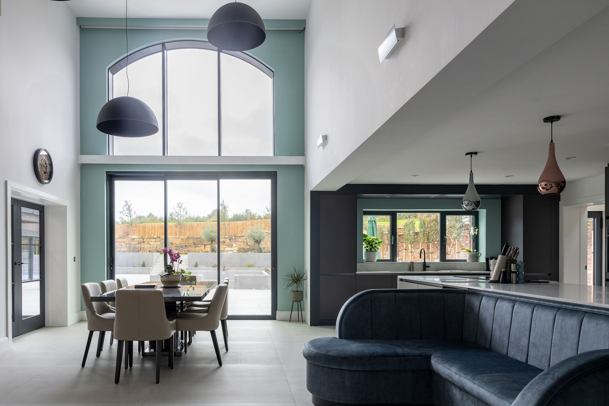 Modern open-plan dining and kitchen area with high arched window, beige chairs around black dining table, blue curved sofa, and pendant lights.