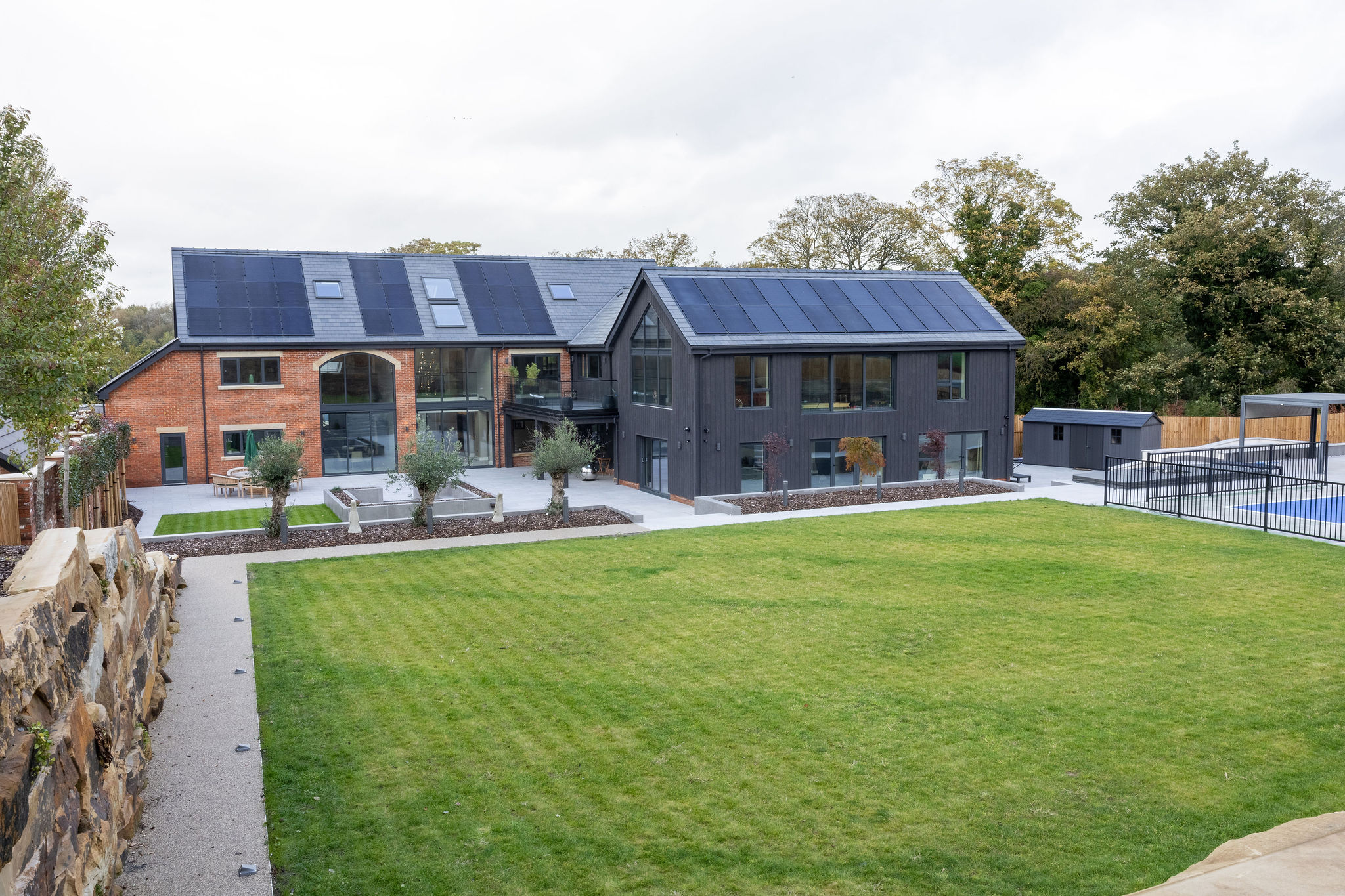 Modern two-story house with red brick and dark wood exterior, solar panels on the roof, large windows, patio, and spacious green lawn.