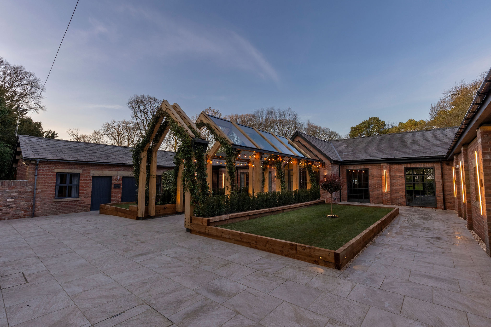 Outdoor courtyard with a glass-roofed wooden pergola adorned with string lights and greenery, surrounded by red brick buildings and paved flooring.