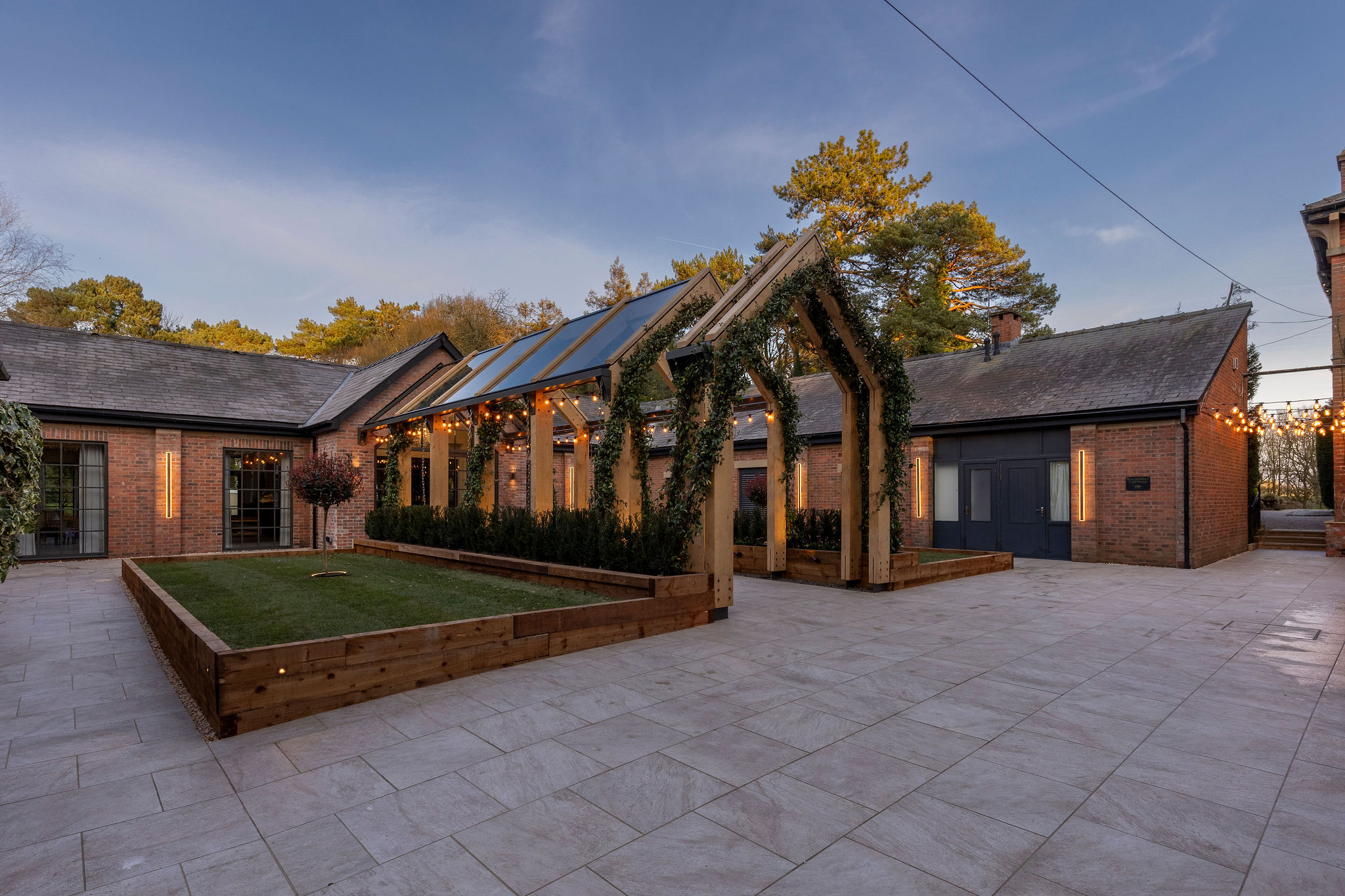 Outdoor patio area with raised grass beds, wooden pergola decorated with greenery and string lights, surrounded by brick buildings at dusk.