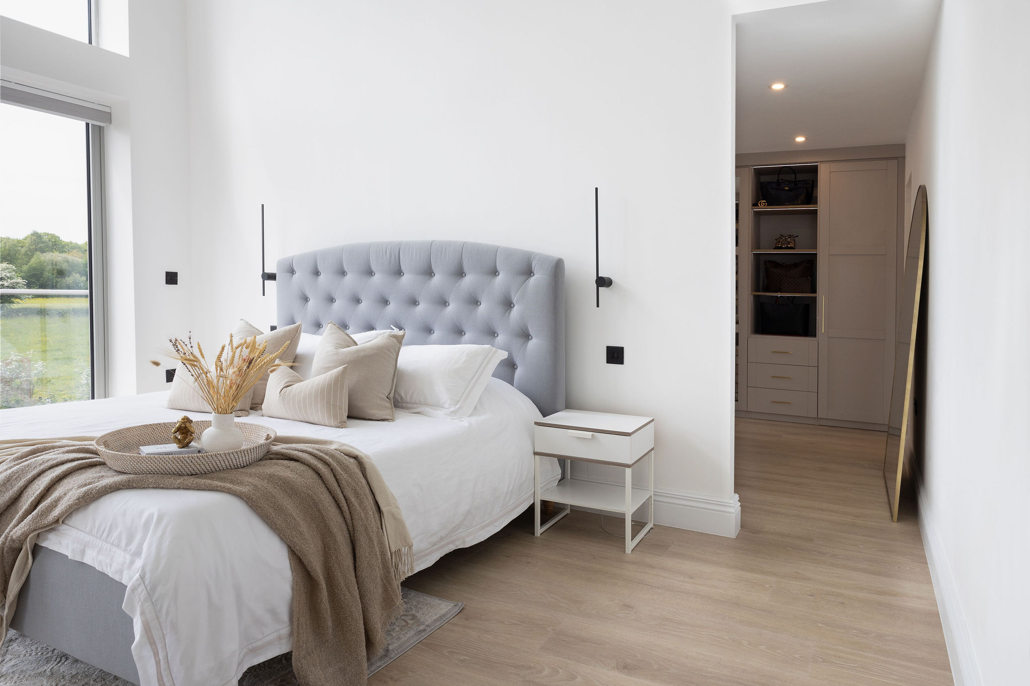 Bright bedroom with a gray upholstered headboard bed, white and beige pillows, beige throw blanket, and a side table next to a walk-in closet.