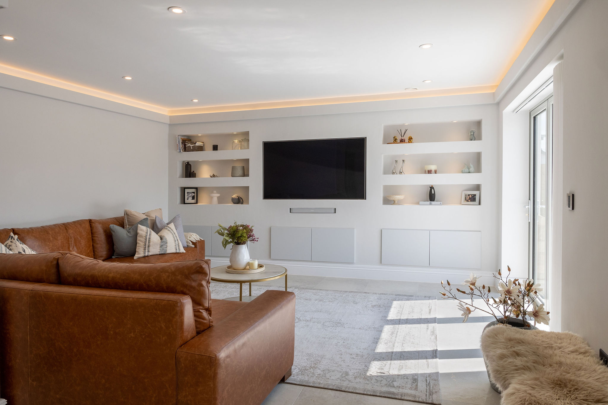 Modern living room with brown leather sofas, built-in shelves, a wall-mounted TV, and a round coffee table with flowers.