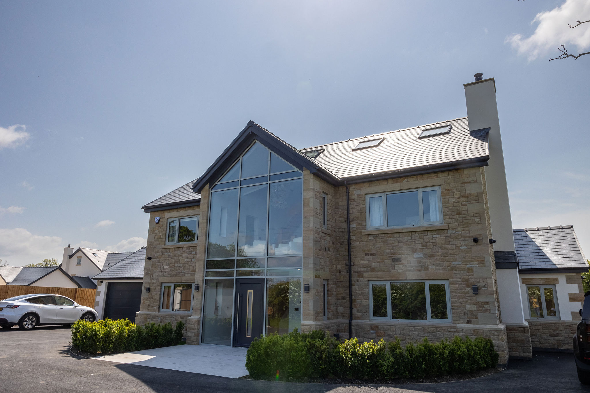 Modern two-story stone house with large glass front entrance, multiple windows, and a black front door under a clear blue sky.