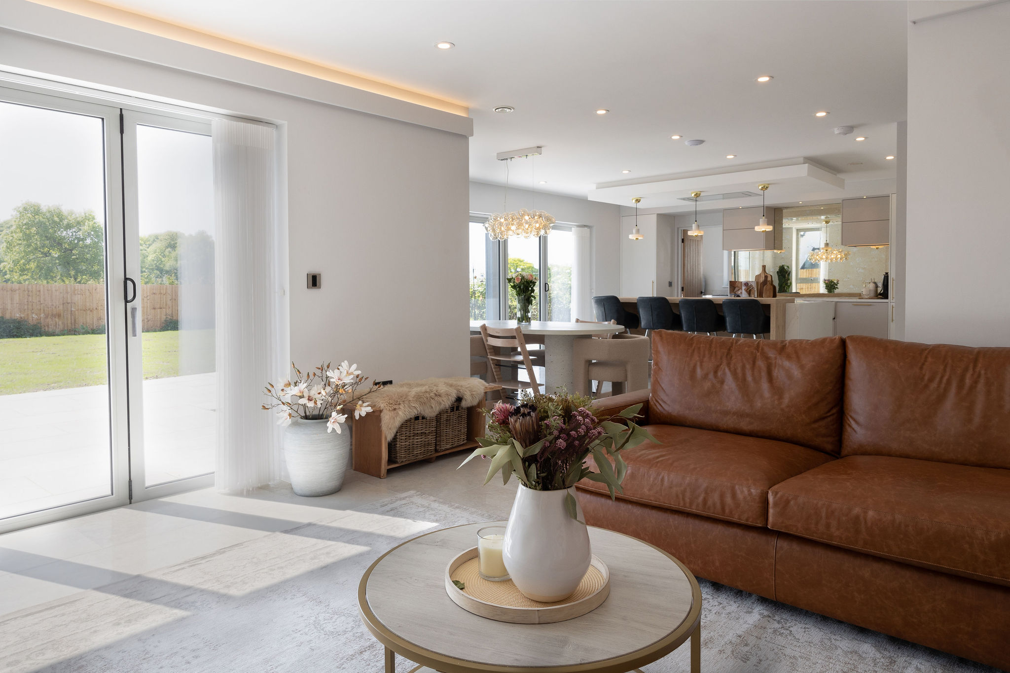 Bright modern living room with brown leather sofa, round coffee table with flower vase, and large sliding glass doors.