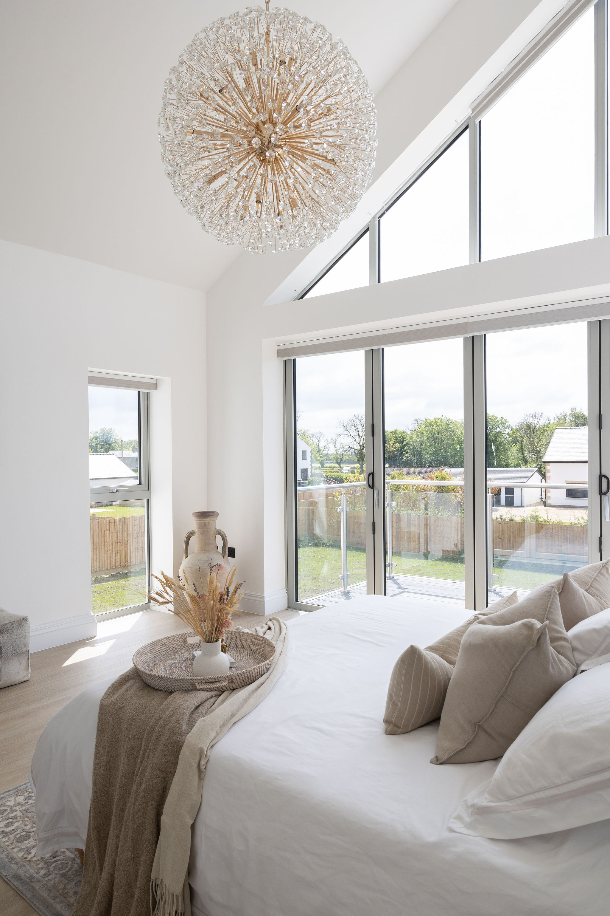 Bright bedroom with large glass sliding doors, a white bed with beige pillows and throw, decorative vase with dried flowers, and a modern spherical chandelier.