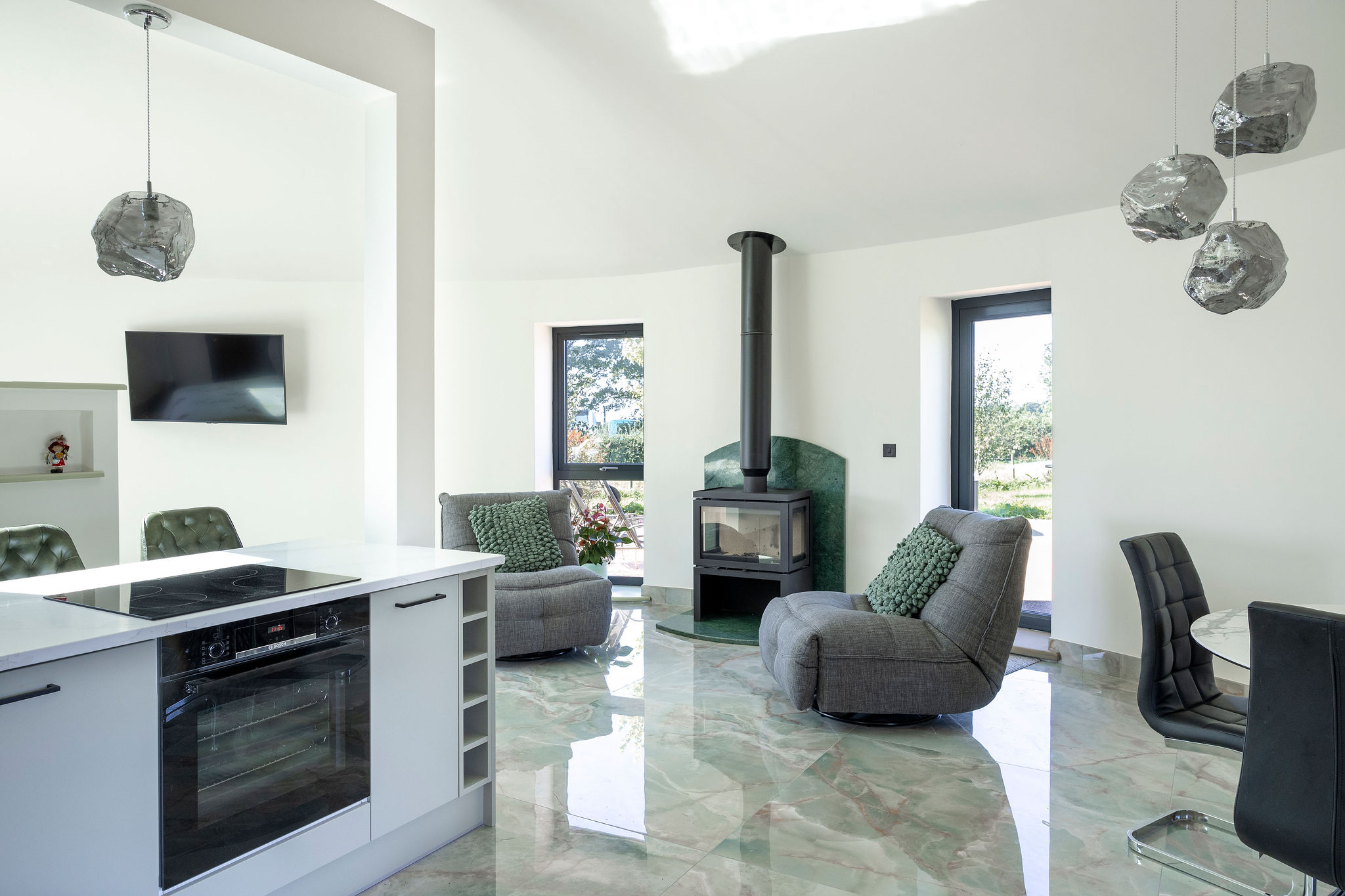 Modern living room with gray armchairs, a black wood stove on a green marble hearth, and a kitchen island with stove and oven.