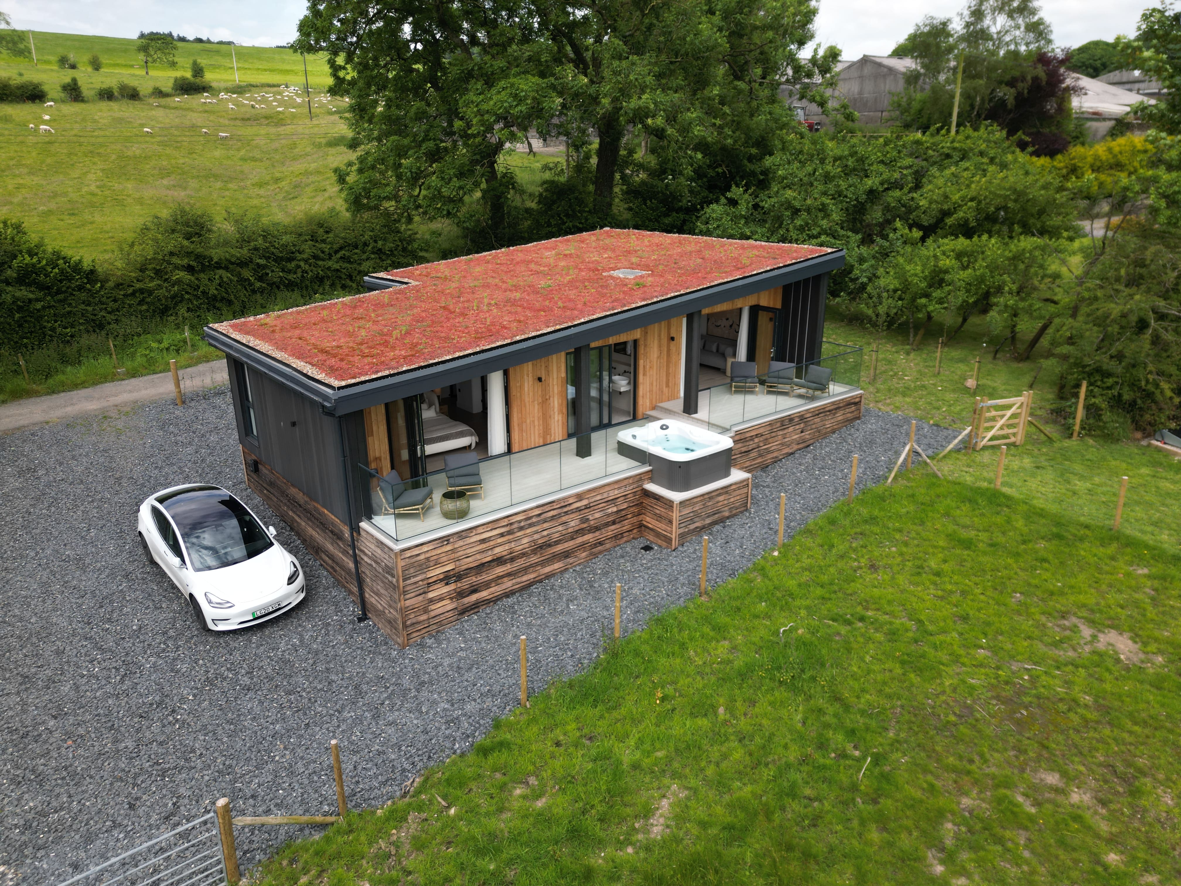 Modern single-story house with a red green roof, wooden and black siding, a glass railing balcony with seating, a hot tub, and a white electric car parked on gravel beside it.