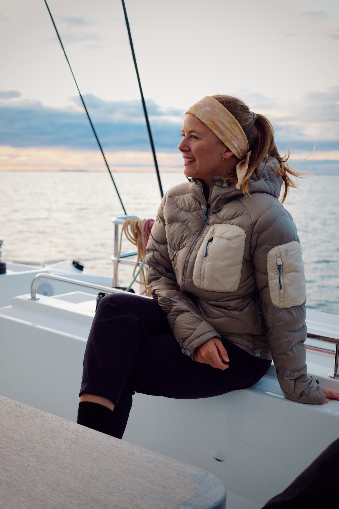 Woman wearing a puffer jacket and headband sitting on a boat railing with a calm sea and cloudy sky in the background.