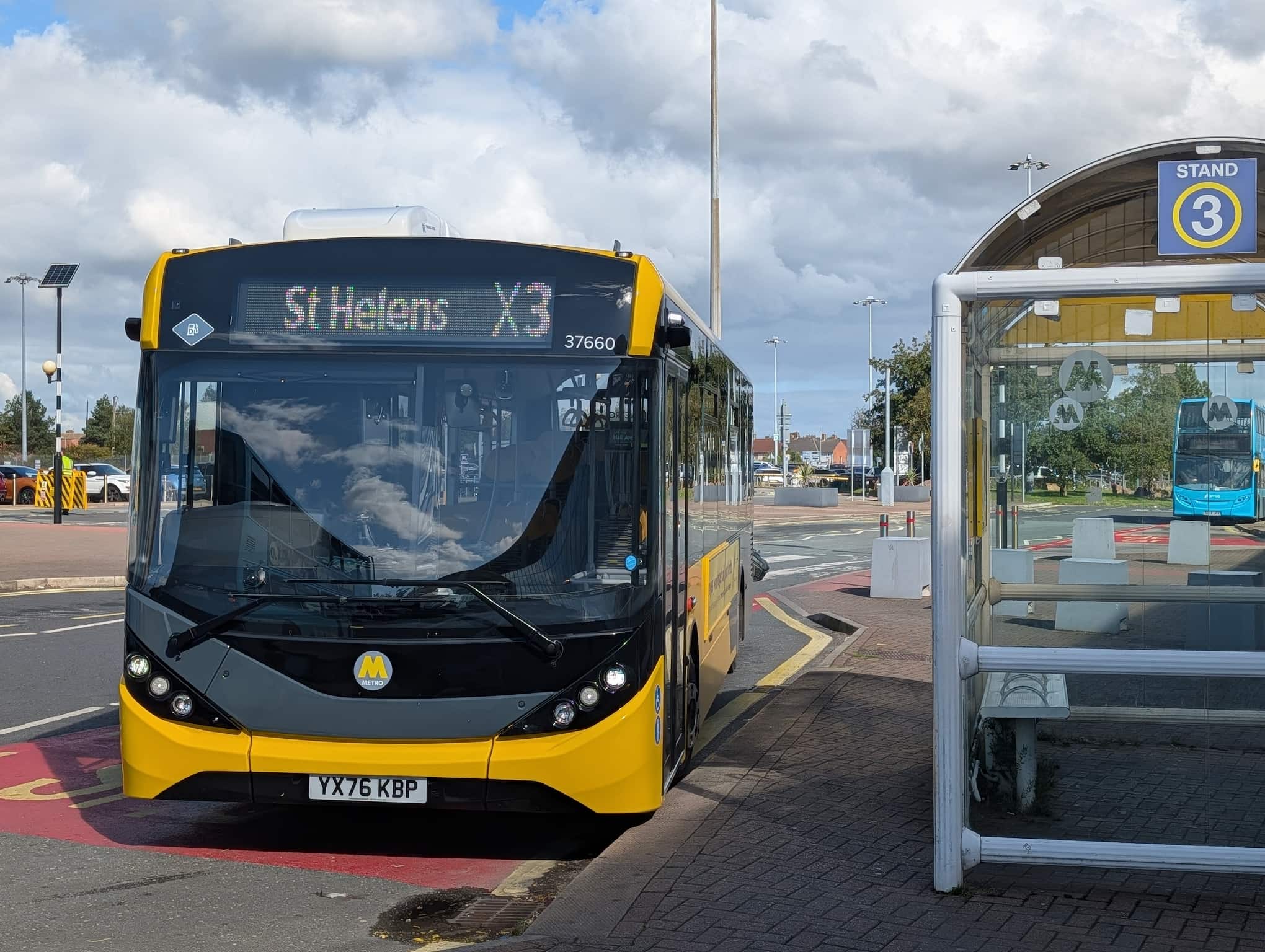 Yellow and black Metro bus with route X3 to St Helens parked at bus stand 3 next to a glass shelter with a bench on a cloudy day.
