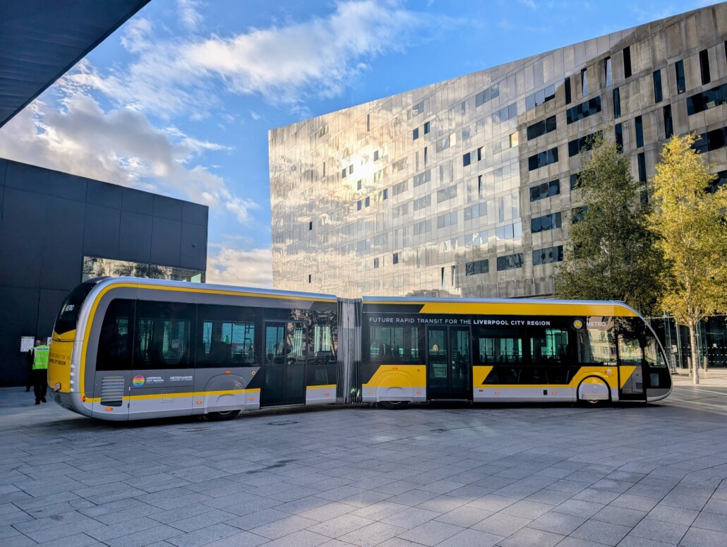 Yellow and black articulated bus parked in an urban plaza with modern glass and concrete buildings and trees in the background under a partly cloudy blue sky.