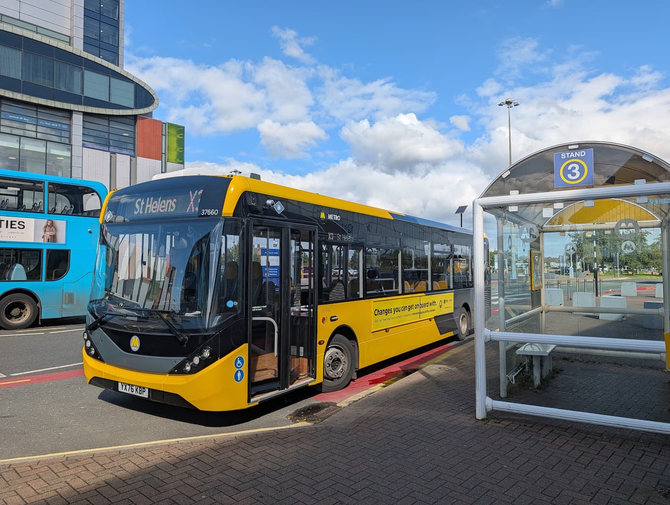 Yellow and black metro bus labeled X3 to St Helens parked at bus stand 3 with a glass bus shelter on the right and a blue double-decker bus in the background.