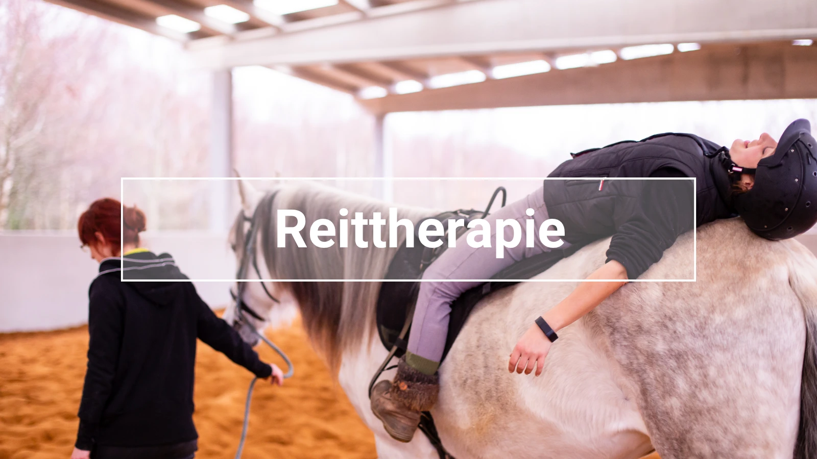 Weißes Pferd mit grauer Mähne wird in einer Halle geführt, trägt ein Sattelpad und Reitpad; eine Frau mit Reithelm liegt entspannt rücklings auf dem Pferd – typische Szene aus der Reittherapie.