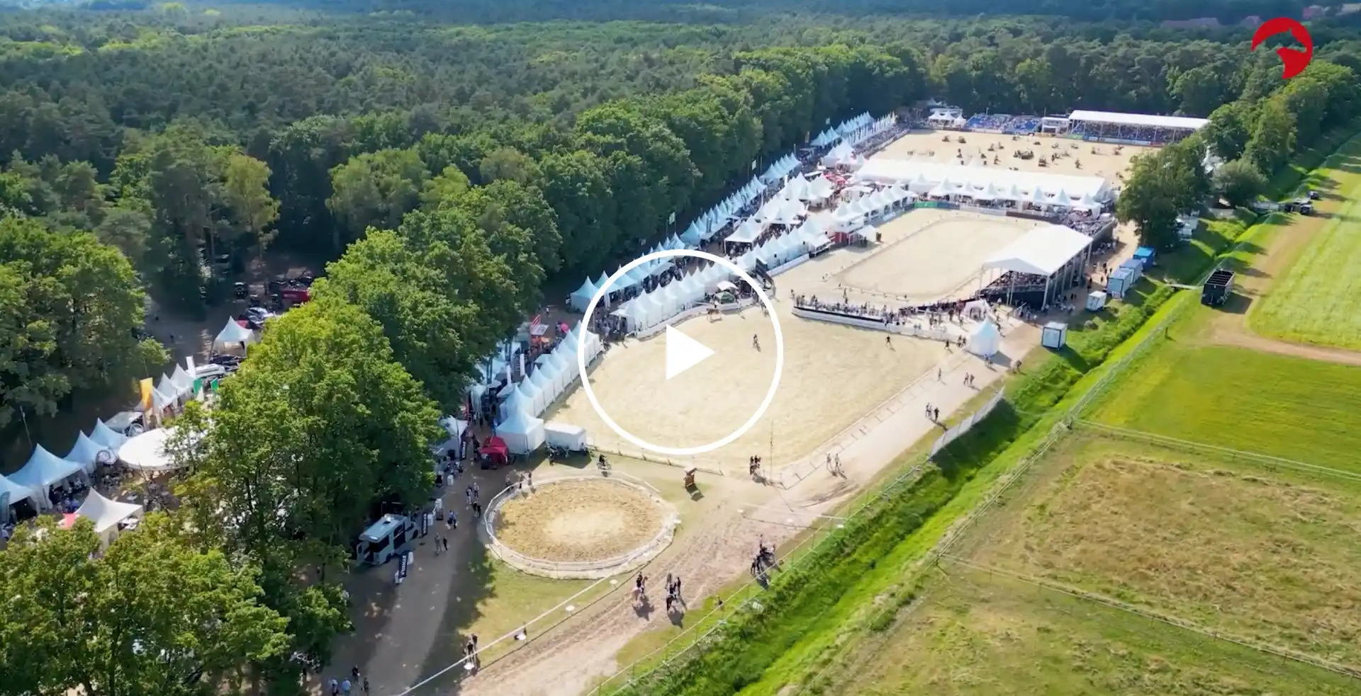 Luftaufnahme einer Reitsportveranstaltung mit mehreren Reitplätzen, weißen Zelten und Zuschauertribünen am Waldrand.Video-Button in der Mitte.