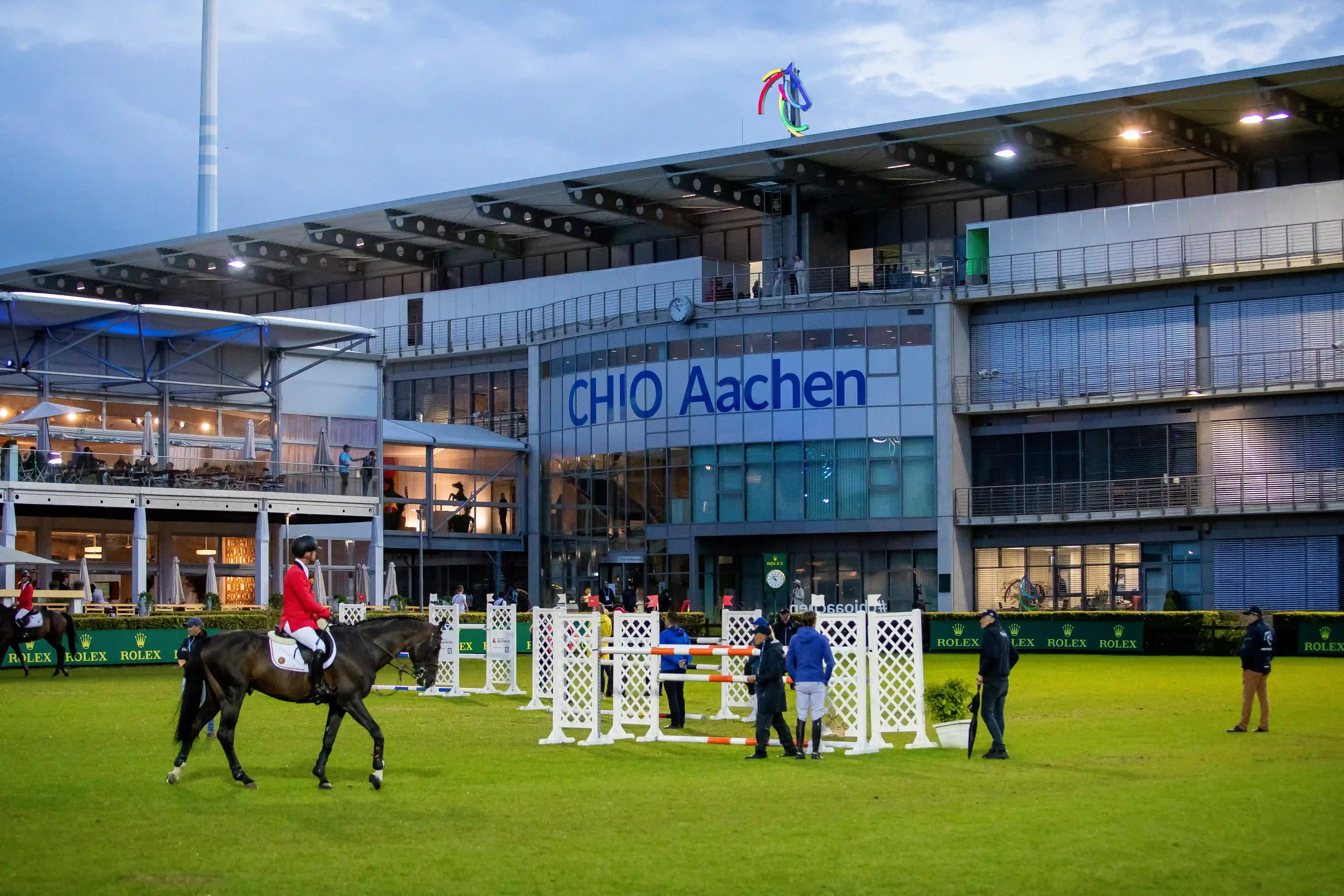 Abendstimmung beim CHIO Aachen – Reiter und Pferde beim Platztraining vor dem Hauptgebäude des Turniergeländes mit Springhindernissen und Rolex-Werbebanden