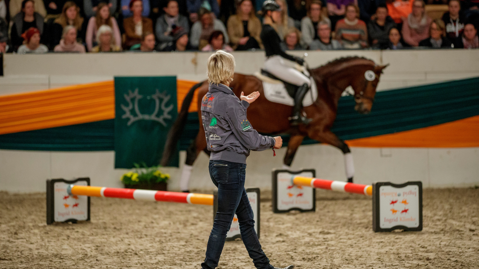 Ausbilderin in einer Reithalle gibt Anweisungen, im Hintergrund reitet eine Person ein Pferd vor Publikum