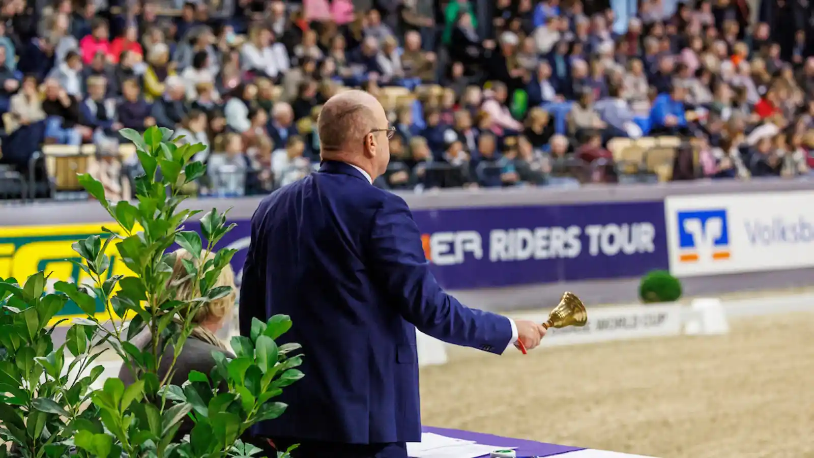 Richter in einer Reithalle, von hinten zu sehen, hält eine Glocke in der Hand; im Hintergrund sitzen Zuschauer auf der Tribüne und Bandenwerbung ist sichtbar.
