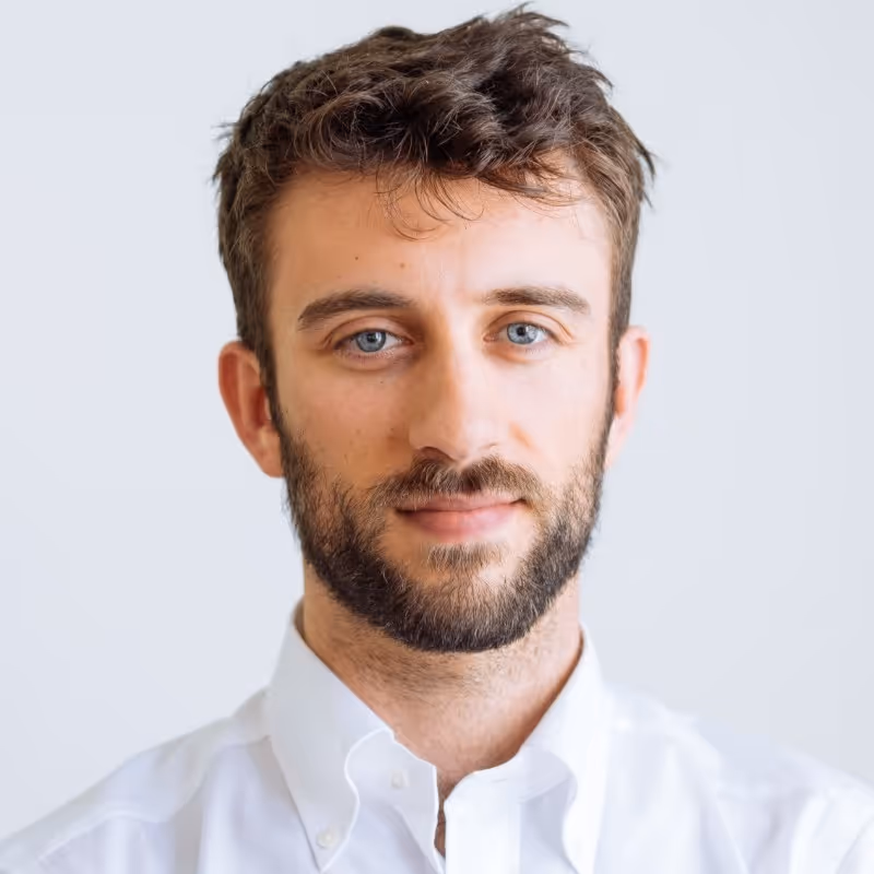 Portrait of a young man with blue eyes, beard, and curly brown hair wearing a white button-up shirt against a plain white background.