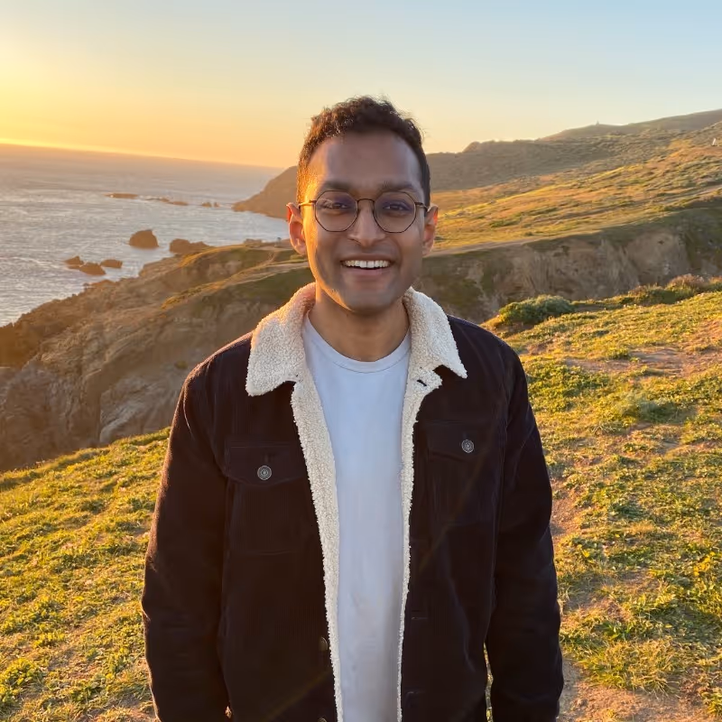 Smiling man wearing glasses and a dark jacket with white fleece lining standing on a grassy hillside at sunset with ocean and rocky cliffs in the background.