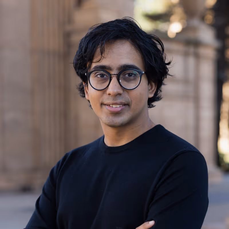 Man with black glasses and black sweater smiling outdoors near stone columns.