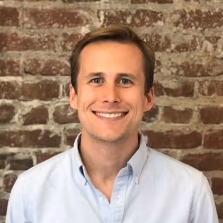 Smiling young man with light brown hair wearing a light blue button-up shirt standing in front of a brick wall.