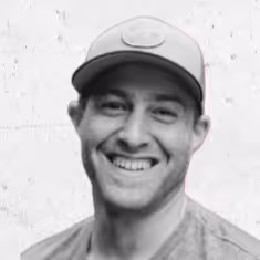 Smiling man wearing a baseball cap and a casual shirt against a light textured wall.