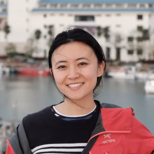 Smiling young woman with black hair wearing a red Arc'teryx jacket and a black sweater with white stripes, standing outdoors near water with buildings in the background.