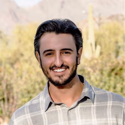 Smiling man with short dark hair and beard wearing a gray plaid shirt outdoors with greenery and mountains in the background.