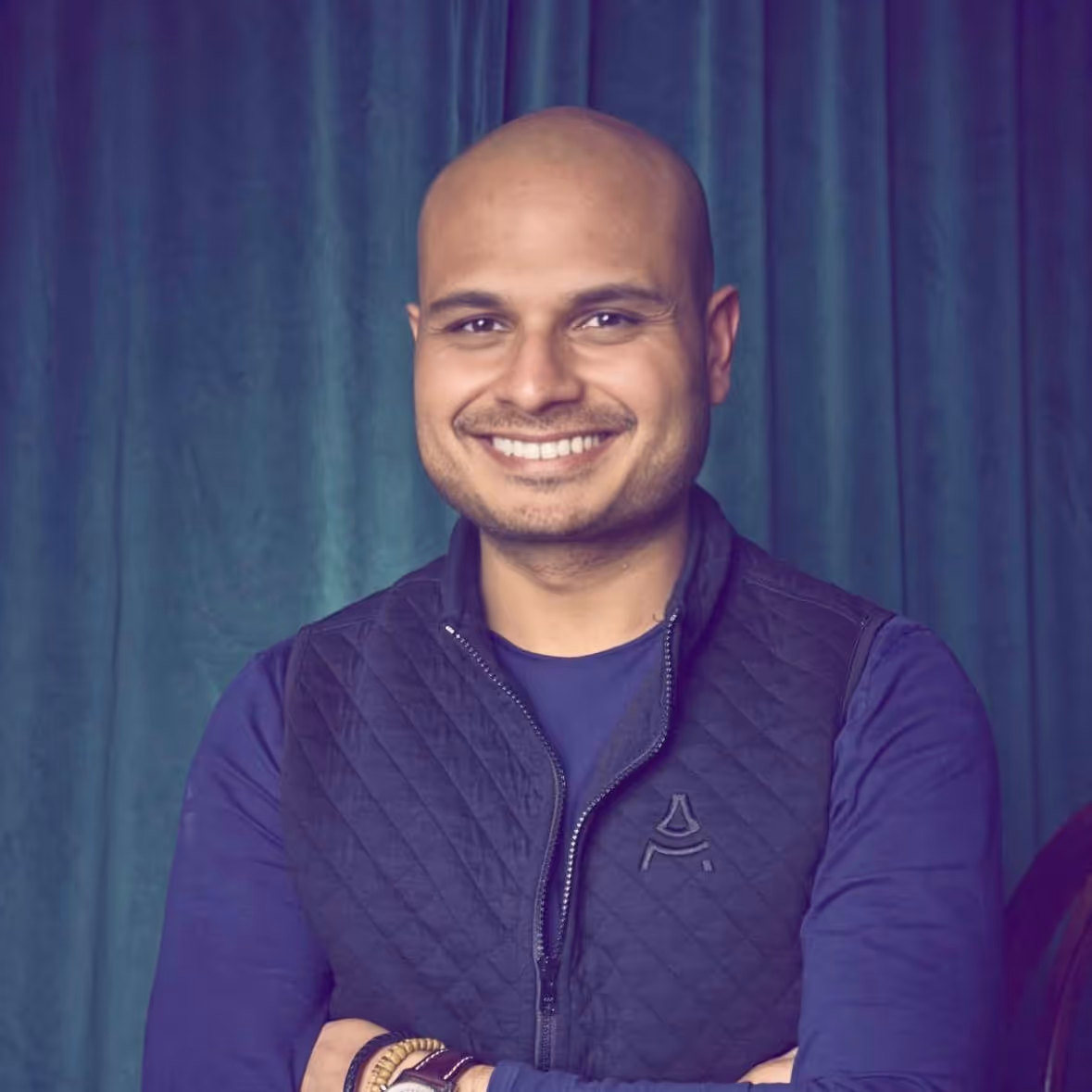 Smiling bald man with crossed arms wearing a navy blue quilted vest and long-sleeve shirt, standing against a teal curtain backdrop.