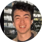 Young man with dark curly hair smiling in front of a window with vertical blinds.
