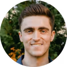 Smiling young man with short brown hair and greenery background.