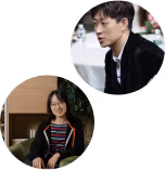 Two young adults; a man in a black jacket looking to the side and a smiling woman with glasses sitting indoors.