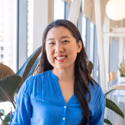 Smiling woman with long dark hair wearing a blue blouse in a bright indoor setting with large windows and plants.