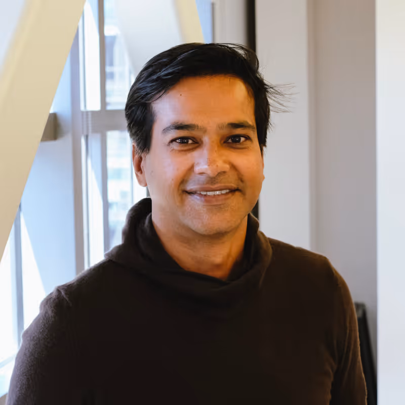 Smiling man with dark hair wearing a dark brown sweater standing indoors near a window.