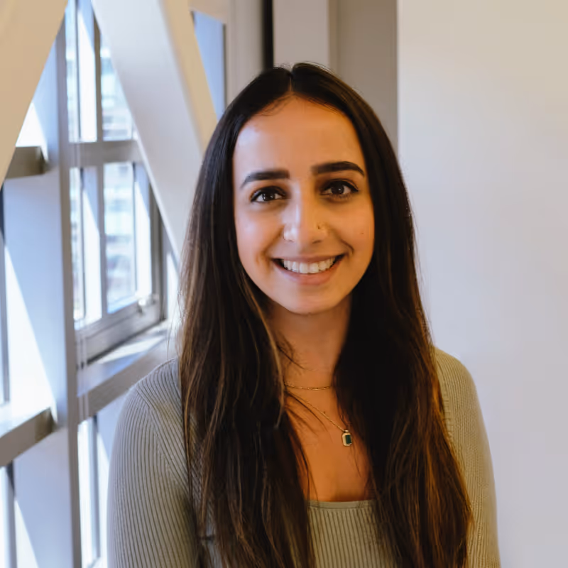 Smiling woman with long dark hair wearing a light gray ribbed top and a green pendant necklace, standing indoors near windows.