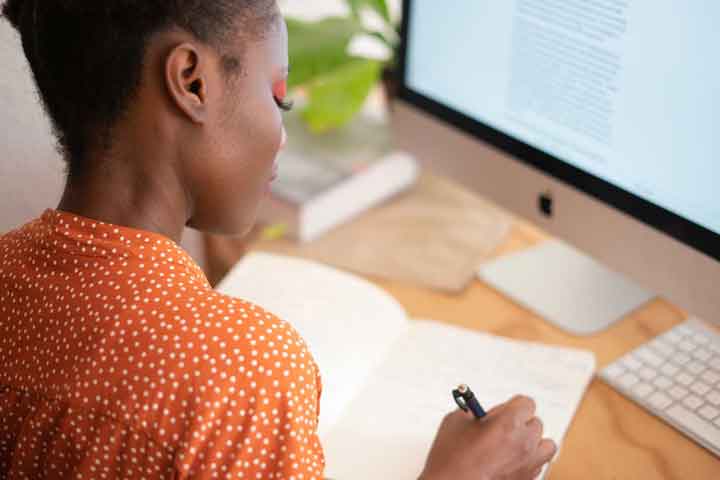 woman sitting at desk writing in notepad, in front of desktop computer