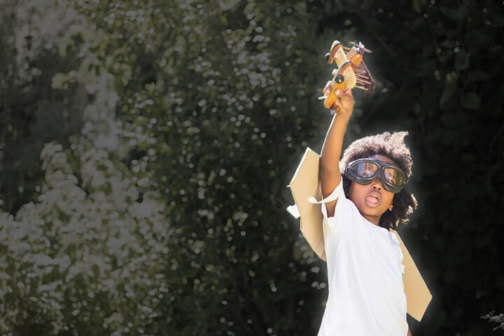 boy standing in field playing with toy airplane, wearing pilot's goggles and wings strapped to his back