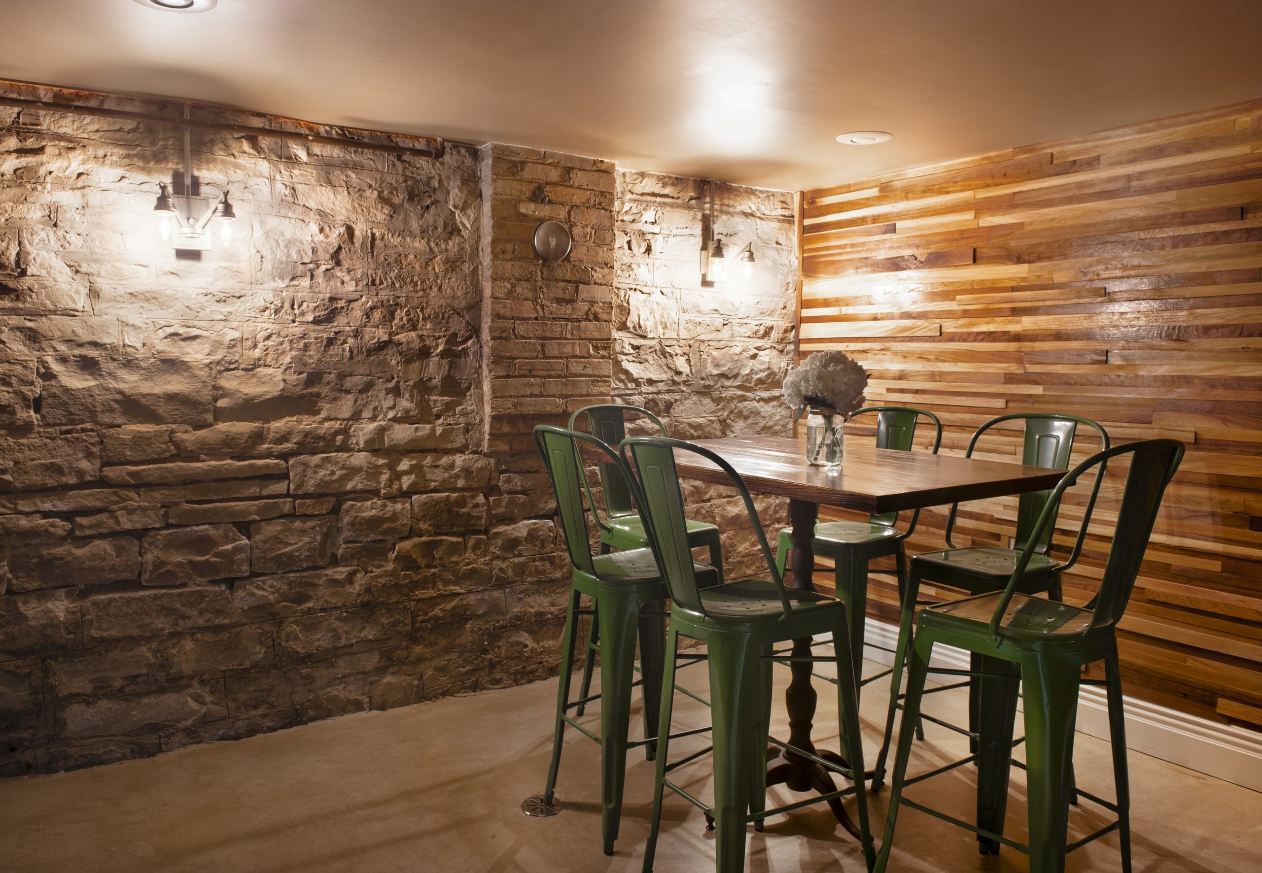 Rustic dining area with a wooden table, six green metal chairs, exposed stone and wood-paneled walls, and a vase of white flowers on the table.